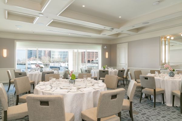 The image shows an elegant banquet room with round tables, each covered with white tablecloths and set for a meal, with floral centerpieces.