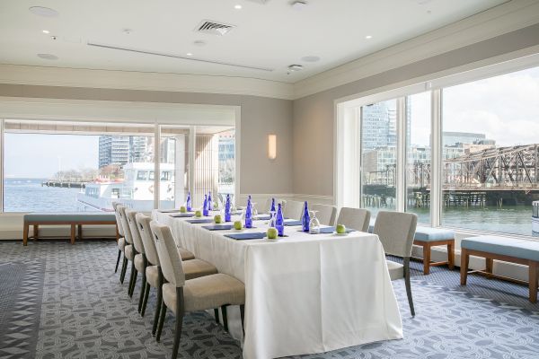 A conference room with a table set for a meeting, overlooking a water view with a docked boat and cityscape in the background.