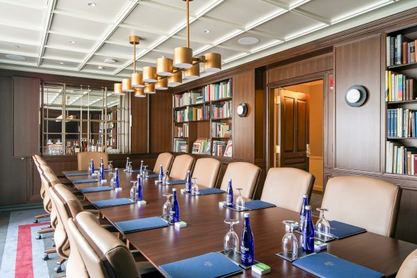 The image shows a conference room with a long table, leather chairs, water bottles, glasses, notepads, and bookshelves.