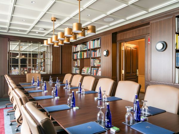 The image shows a conference room with a long table, leather chairs, water bottles, glasses, notepads, and bookshelves.