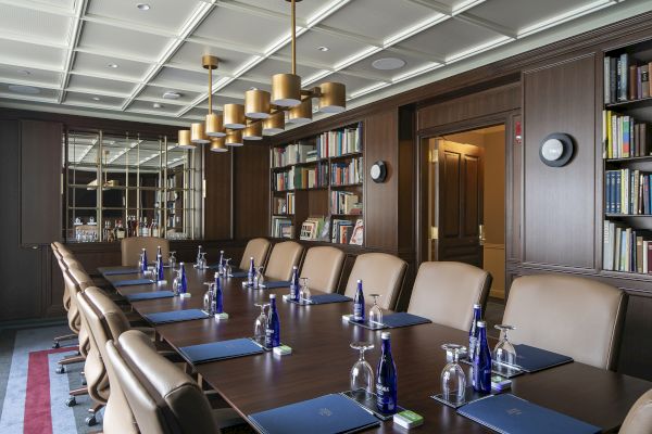 A conference room with a long table, leather chairs, water bottles, glasses, notebooks, a bookshelf, and ceiling lights is shown.