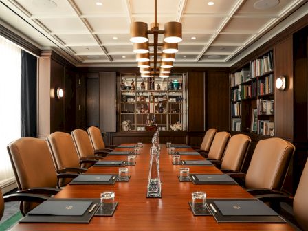 A formal boardroom with a long wooden table, leather chairs, and bookshelves under warm ceiling lights.