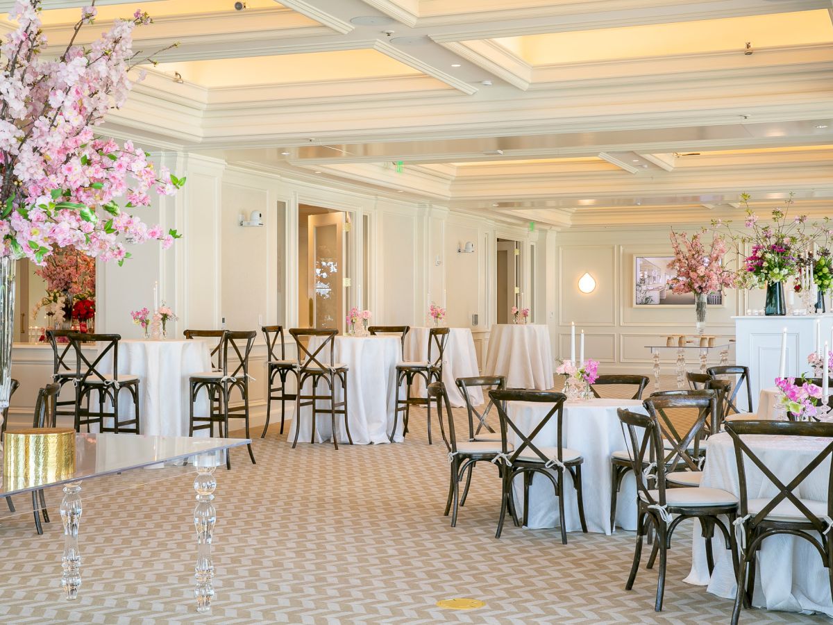 Elegant event space with round tables, chairs, and large floral centerpieces in shades of pink and white, and soft lighting.