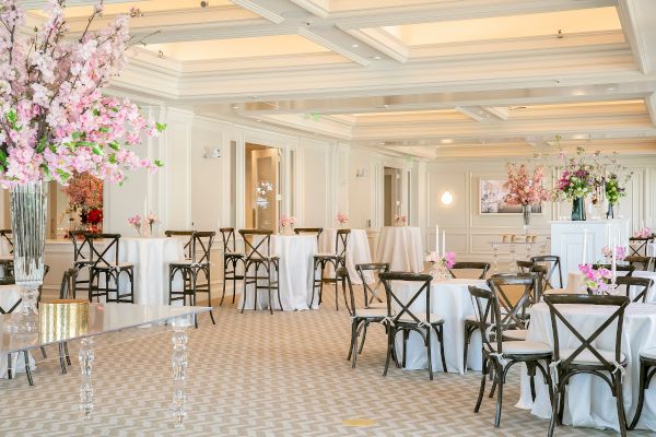 The image shows an elegantly decorated banquet hall with floral arrangements, white tablecloths, wooden chairs, and lit candles on tables.