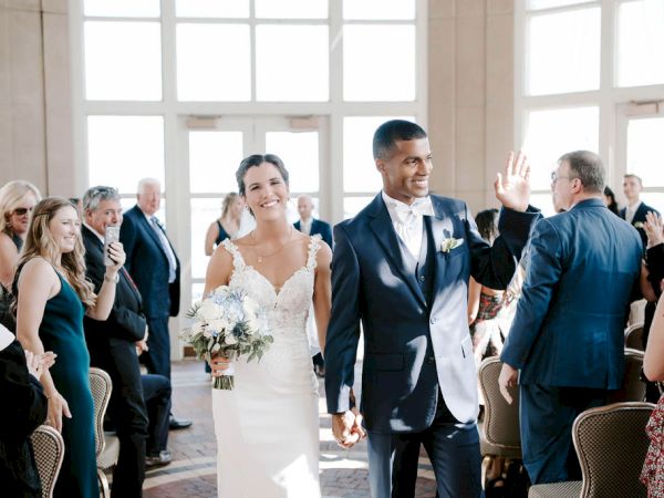 A smiling couple in wedding attire walks down an aisle, greeted by applauding guests in a bright room full of windows.