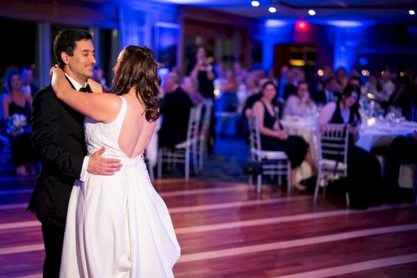 A couple is dancing in a warmly lit room, surrounded by seated guests watching them.