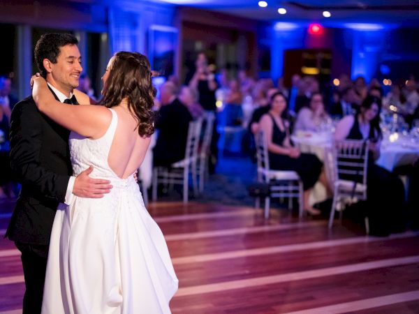 A couple is dancing in a warmly lit room, surrounded by seated guests watching them.