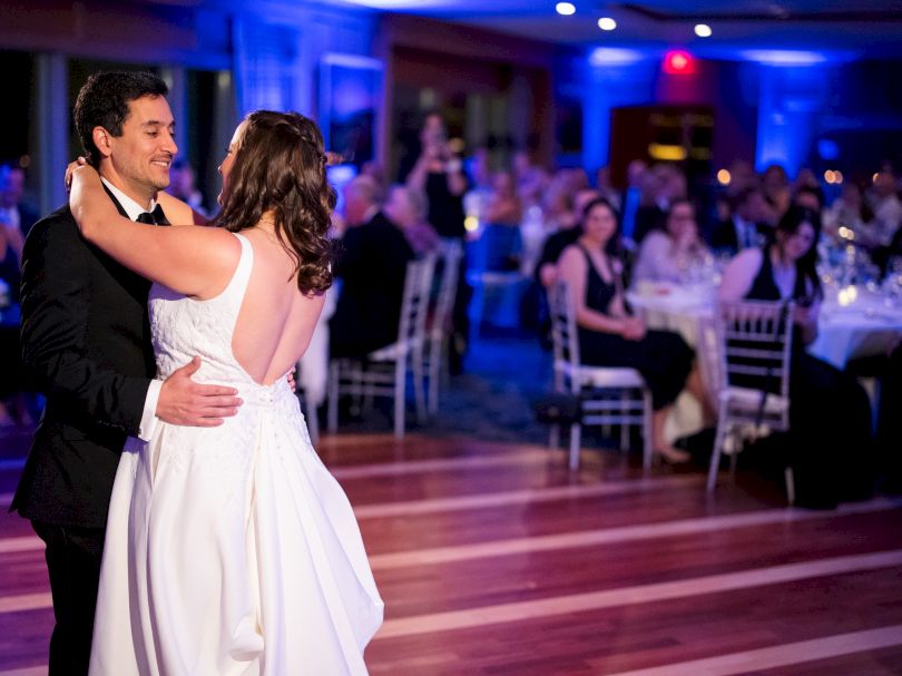 A couple is dancing in a warmly lit room, surrounded by seated guests watching them.