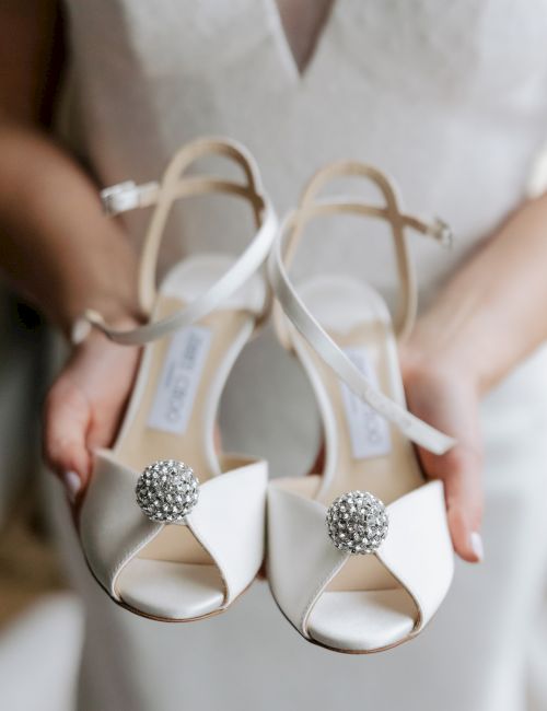 A person is holding a pair of elegant white shoes with jeweled embellishments on the toes.