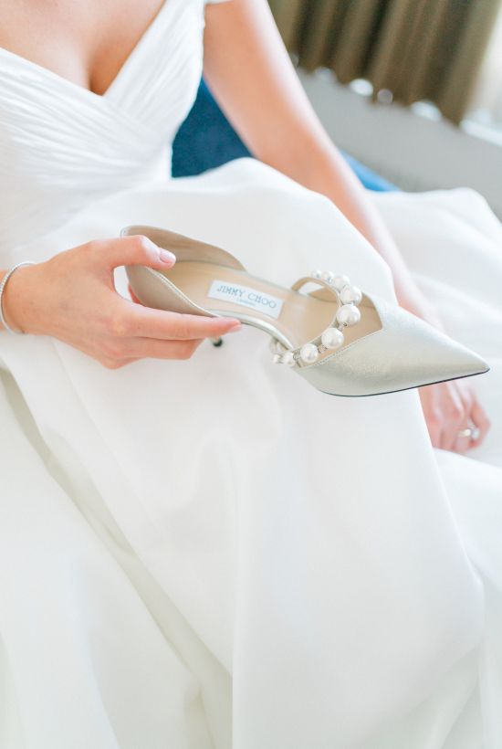 A person in a white dress holds an elegant high-heeled shoe with pearl embellishments, possibly preparing for a special occasion.