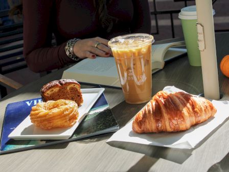 A table with a croissant, pastry, iced coffee, a magazine, and a person reading a book under sunlight.