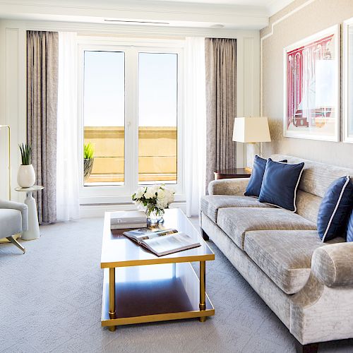 A modern living room with a sofa, armchair, coffee table, lamps, and framed artwork. There are curtains and a door leading to an outdoor area.