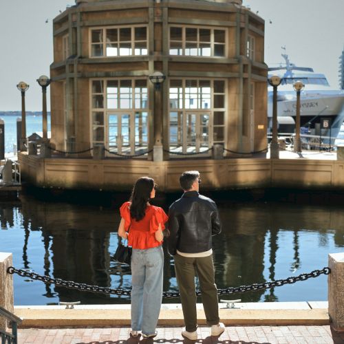 A couple stands by the water, facing a domed building with large windows. A yacht is visible nearby on a sunny day.