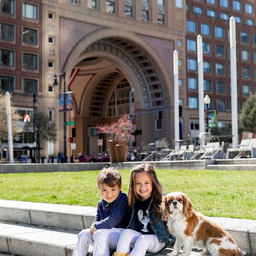 Two children and a dog sit on steps in a city park, with a large archway and buildings in the background.