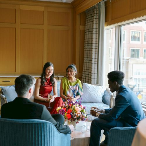 A group of four people sit in a cozy, modern room with wooden panels, engaged in conversation around a table with flowers.