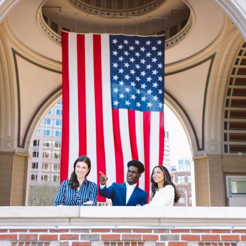 Three people stand on a brick balcony beneath a large American flag, with city buildings visible in the background.