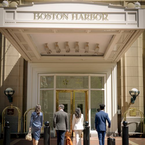 Four people with luggage walk toward the entrance of Boston Harbor Hotel, a luxurious building with ornate architecture.