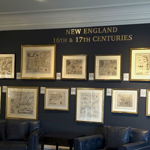 The image shows framed maps on a wall labeled "New England 16th & 17th Centuries" with dark seating below.