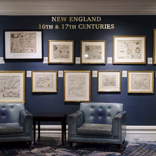 The image shows a display of framed maps and documents titled "New England 16th & 17th Centuries" above two chairs and a table.
