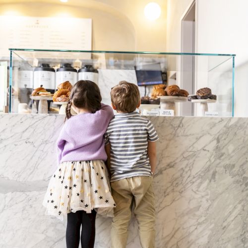 Two children are standing at a marble bakery counter, looking at a display of pastries under a glass case.