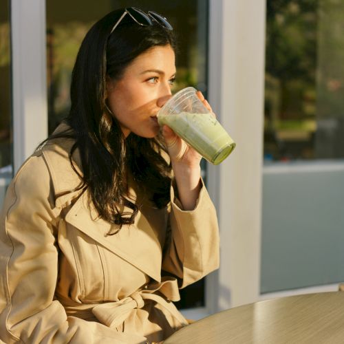 A person in a beige coat is drinking a green beverage while sitting at an outdoor table in the sunlight.