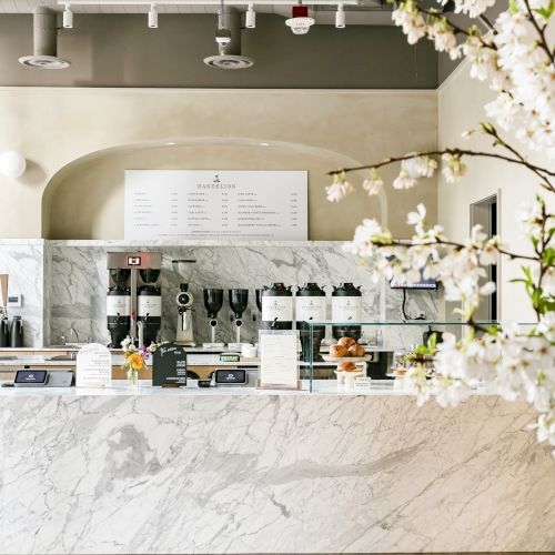 A modern café interior featuring a marble counter, coffee equipment, menu board, and decorative flowers in a vase.