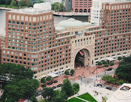 The image shows an archway building by the waterfront with people walking around and trees surrounding it.