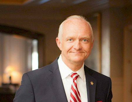 A man in a suit and red striped tie stands indoors, smiling slightly, with his hands behind his back, in a well-lit room with a blurred background.