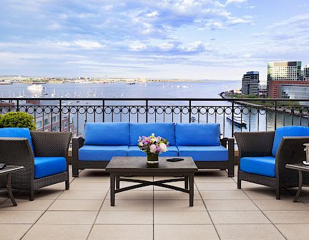 A patio with blue cushioned furniture set facing a scenic waterfront view and city buildings, with a table holding flowers between the seating.