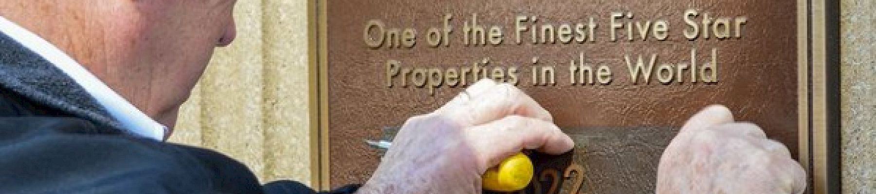 A person is scratching or cleaning a "Forbes" travel guide plaque that reads "One of the Finest Five Star Properties in the World" on a wall.
