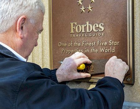 A person is scratching or cleaning a "Forbes" travel guide plaque that reads "One of the Finest Five Star Properties in the World" on a wall.