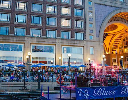 People are gathered at an outdoor venue with umbrellas and seating by a waterfront, near a building and a barge labeled "Blues Barge."