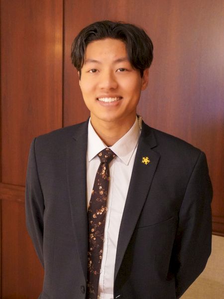 A person in a suit stands indoors, smiling with hands behind their back against a wooden panel background.