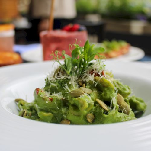 A dish of pasta topped with a creamy green sauce, garnished with seeds and herbs, is served in a white bowl with blurred food in the background.