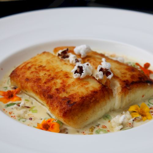 A beautifully plated dish featuring a seared piece of fish, garnished with popcorn and edible flowers, in a creamy sauce.
