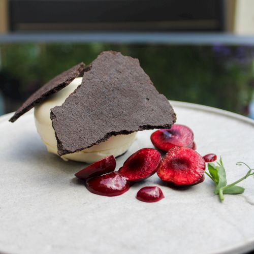 A gourmet dessert with chocolate shards, a cream sphere, sliced cherries, and a garnish, served on a round plate.