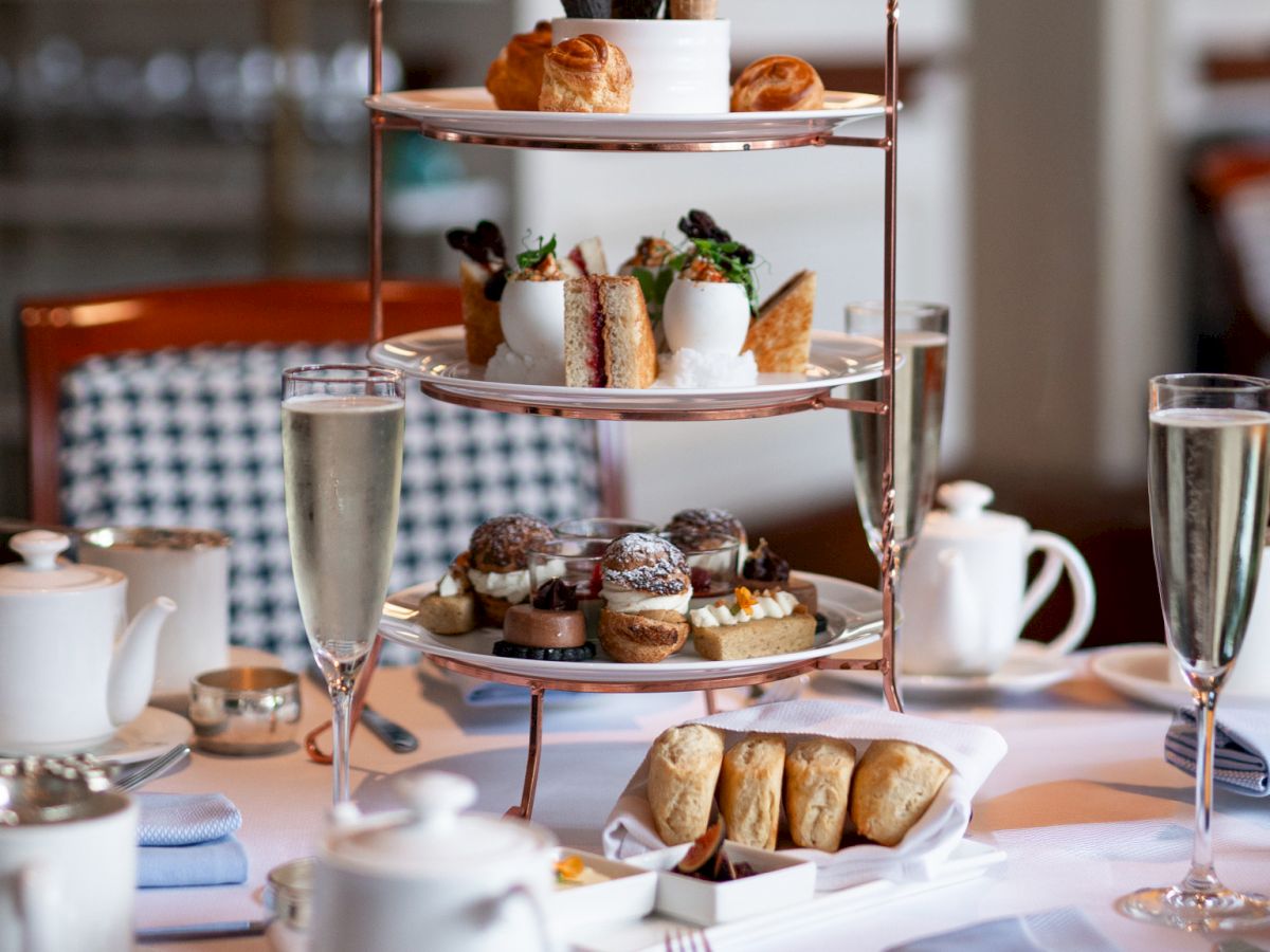 A three-tiered stand of assorted pastries and sandwiches, flanked by champagne flutes, teapots, and delicate tableware for a refined tea service.