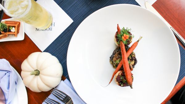 A white plate with a gourmet dish, surrounded by drinks, napkins, pumpkins, and cutlery on a wooden table setting.