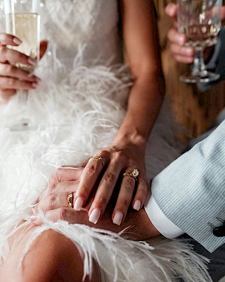 A close-up of a couple's hands with rings, both holding drinks. The person in a white dress appears to be wearing feathers.