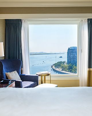 The image shows a hotel room with a cozy chair, lamp, and flowers by a window offering a scenic view of water and buildings outside.