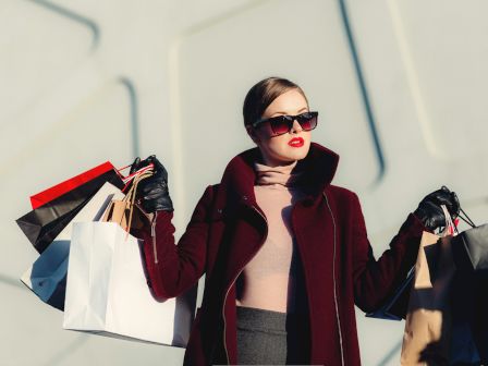 A person wearing sunglasses and a coat holds shopping bags in both hands against a minimalistic background.