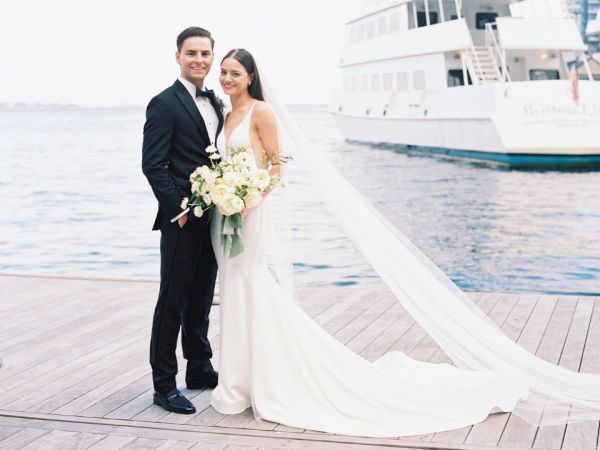 A bride and groom are posing on a dock by the water, with a boat in the background. The bride is holding a bouquet and has a long veil flowing.