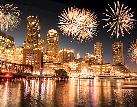 A city skyline at night with fireworks exploding above the harbor and reflected lights on the water.