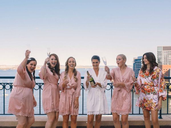 A group of women in robes celebrate with champagne on a terrace overlooking a waterfront cityscape.