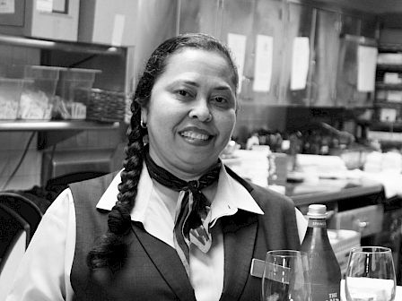 A smiling server in a restaurant holds a tray with a bottle of Mo&euml;t & Chandon, two glasses, and a corkscrew on a napkin, standing in front of the kitchen.