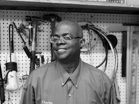 A person wearing a button-up shirt with name embroidery stands in front of a pegboard filled with tools and equipment in a workshop setting.