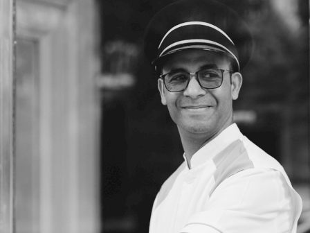 A smiling man wearing a white uniform and cap is holding open a glass door, looking directly at the camera.