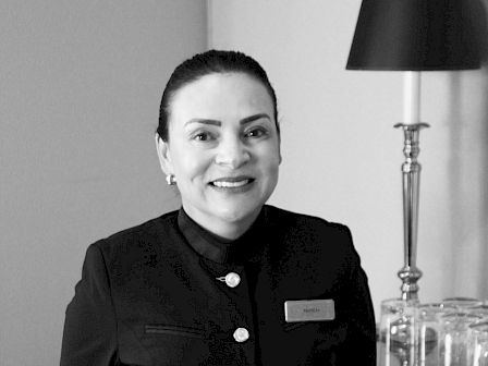 A woman in a uniform is smiling at the camera. She is standing beside a table with several glasses and a lamp on it.