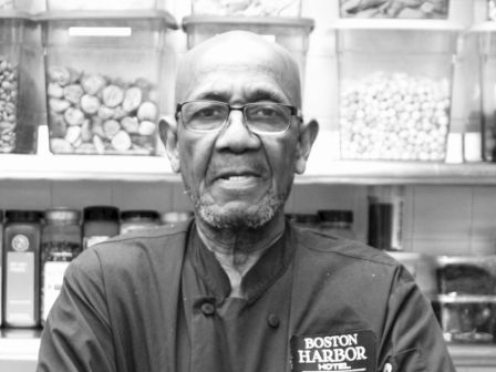 A man in a chef's coat labeled "Boston Harbor" stands with arms crossed, in front of shelves filled with jars of ingredients, facing the camera.