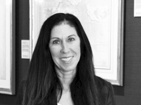 A black-and-white photo of a smiling woman with long hair, standing in front of framed artwork on the wall, dressed in a blazer and white top.
